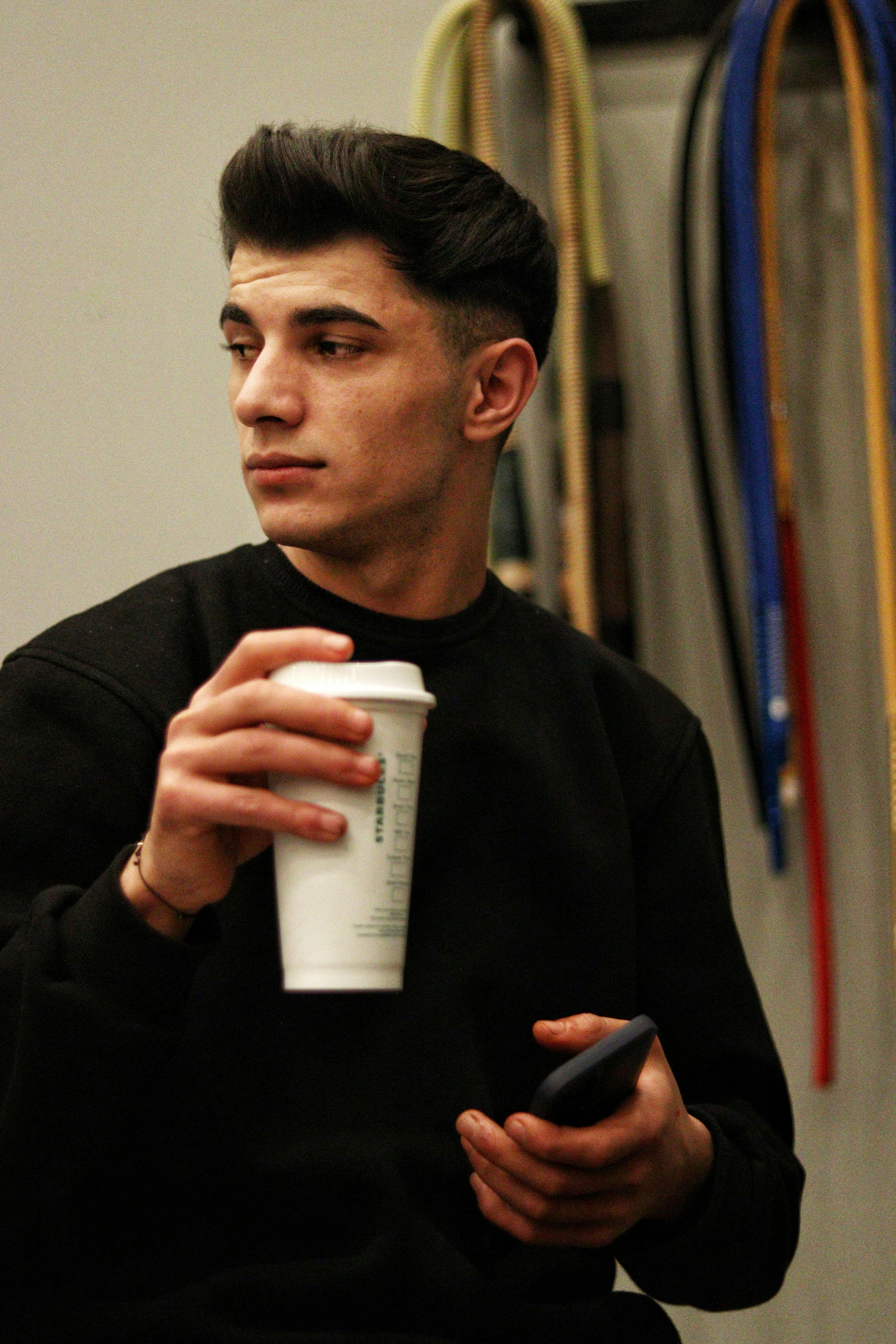 Young man in casual wear holding a coffee cup and smartphone indoors, looking aside.