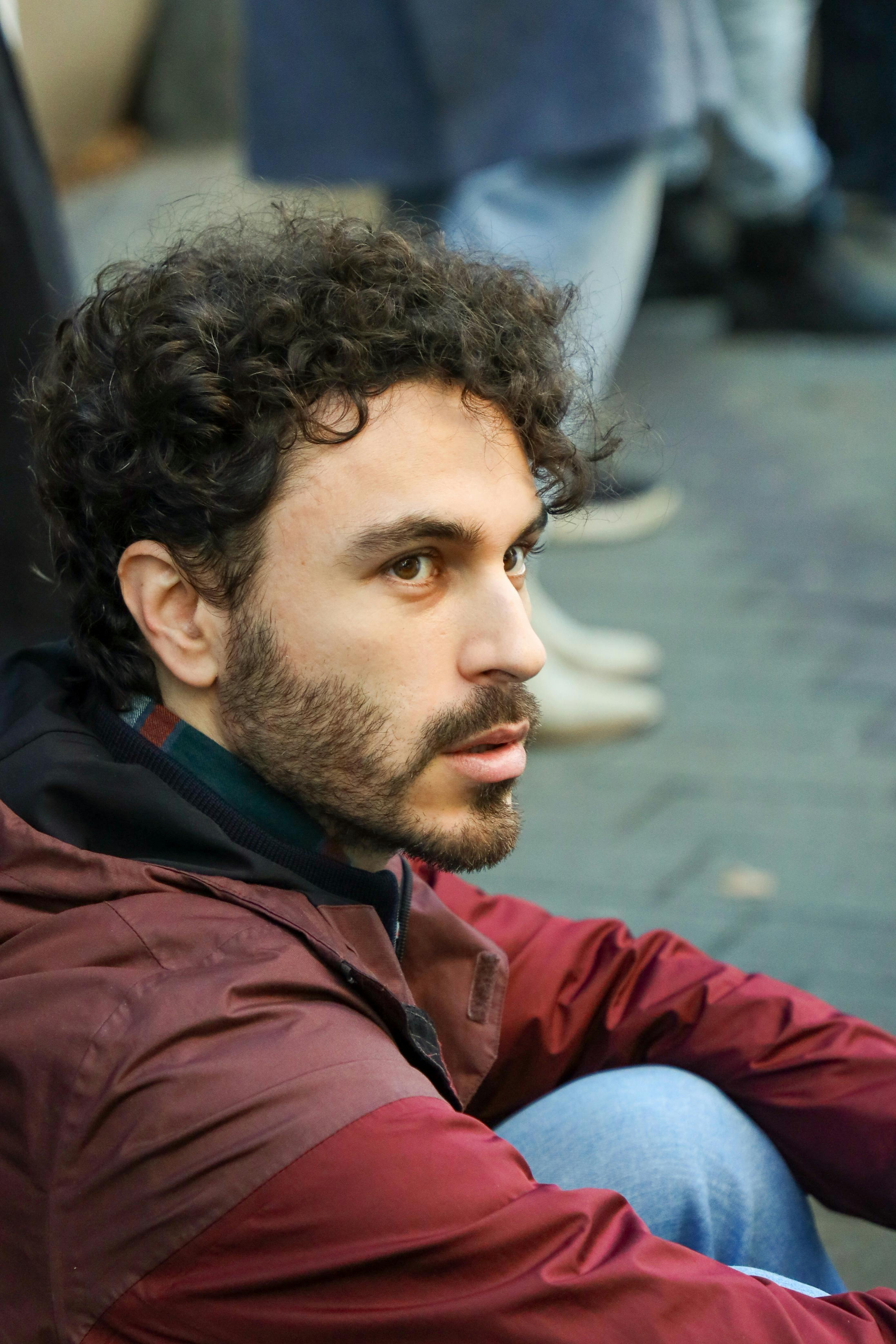 Portrait of a bearded man sitting outdoors, wearing a maroon jacket, reflecting in an urban environment.