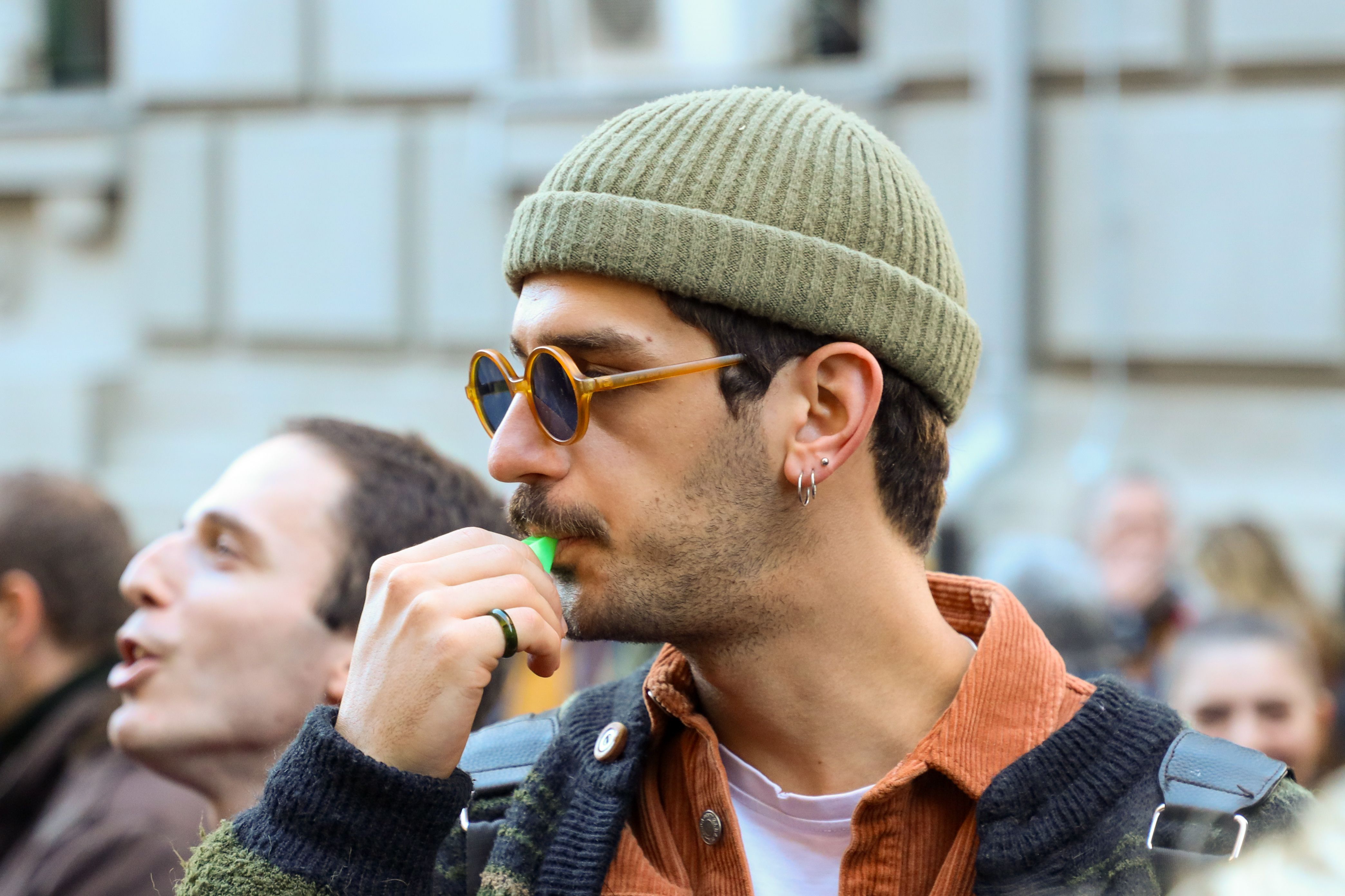 A young man with a beanie and sunglasses whistles during a city protest.