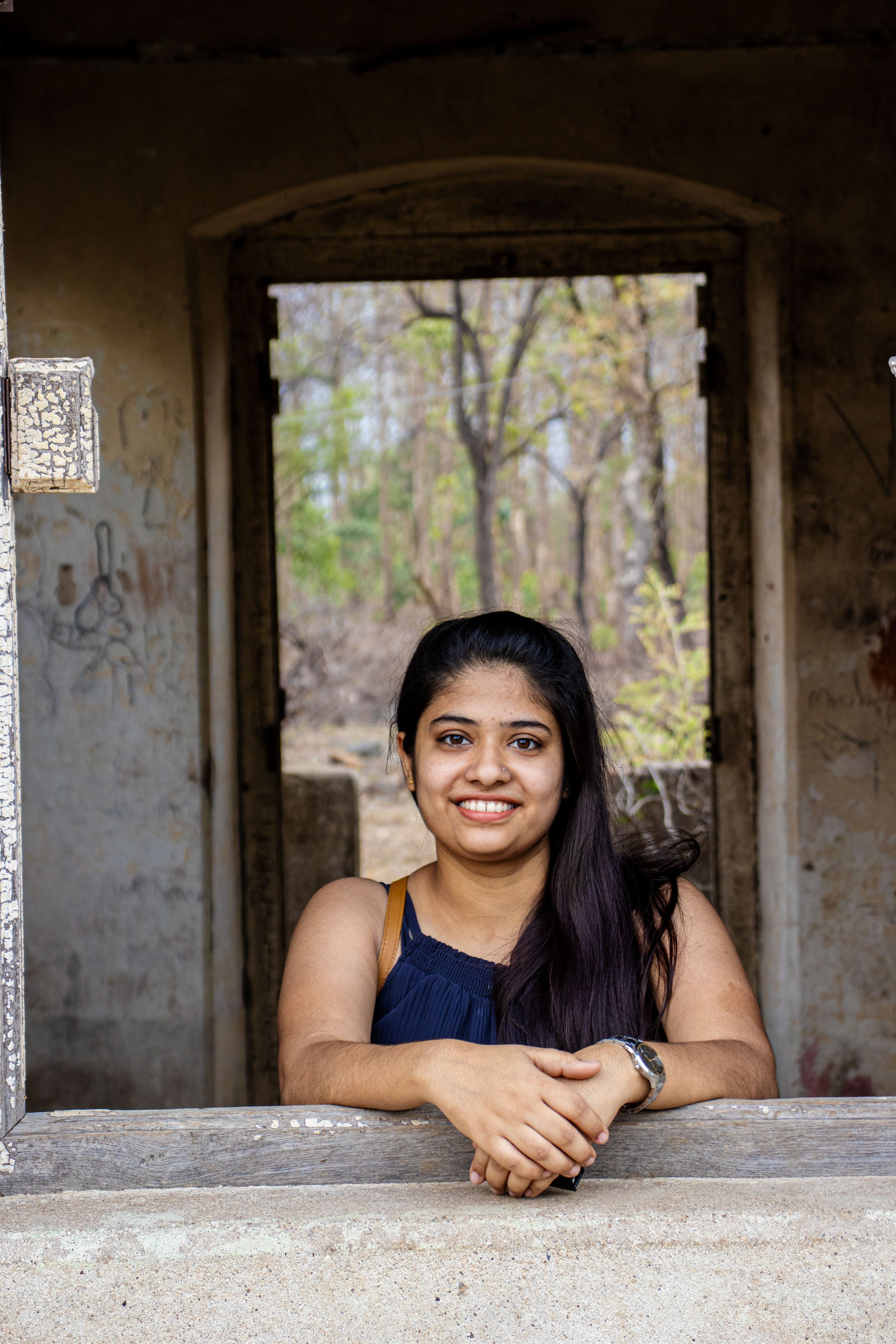 A young woman smiles while posing in an old, abandoned building with a forest view.