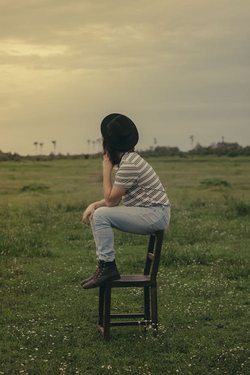 Side view of unrecognizable young female in stylish outfit and hat sitting on wooden chair in peaceful green meadow during sunset