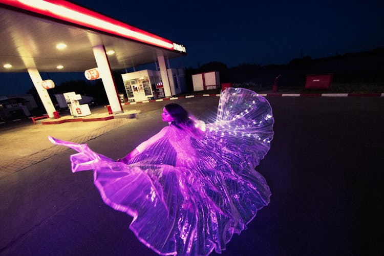 Woman In Glowing Costume Dancing At Gas Station At Night