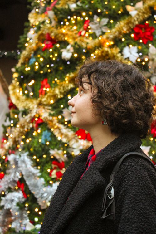 Free A woman in a black coat stands by a decorated Christmas tree, capturing the holiday spirit. Stock Photo