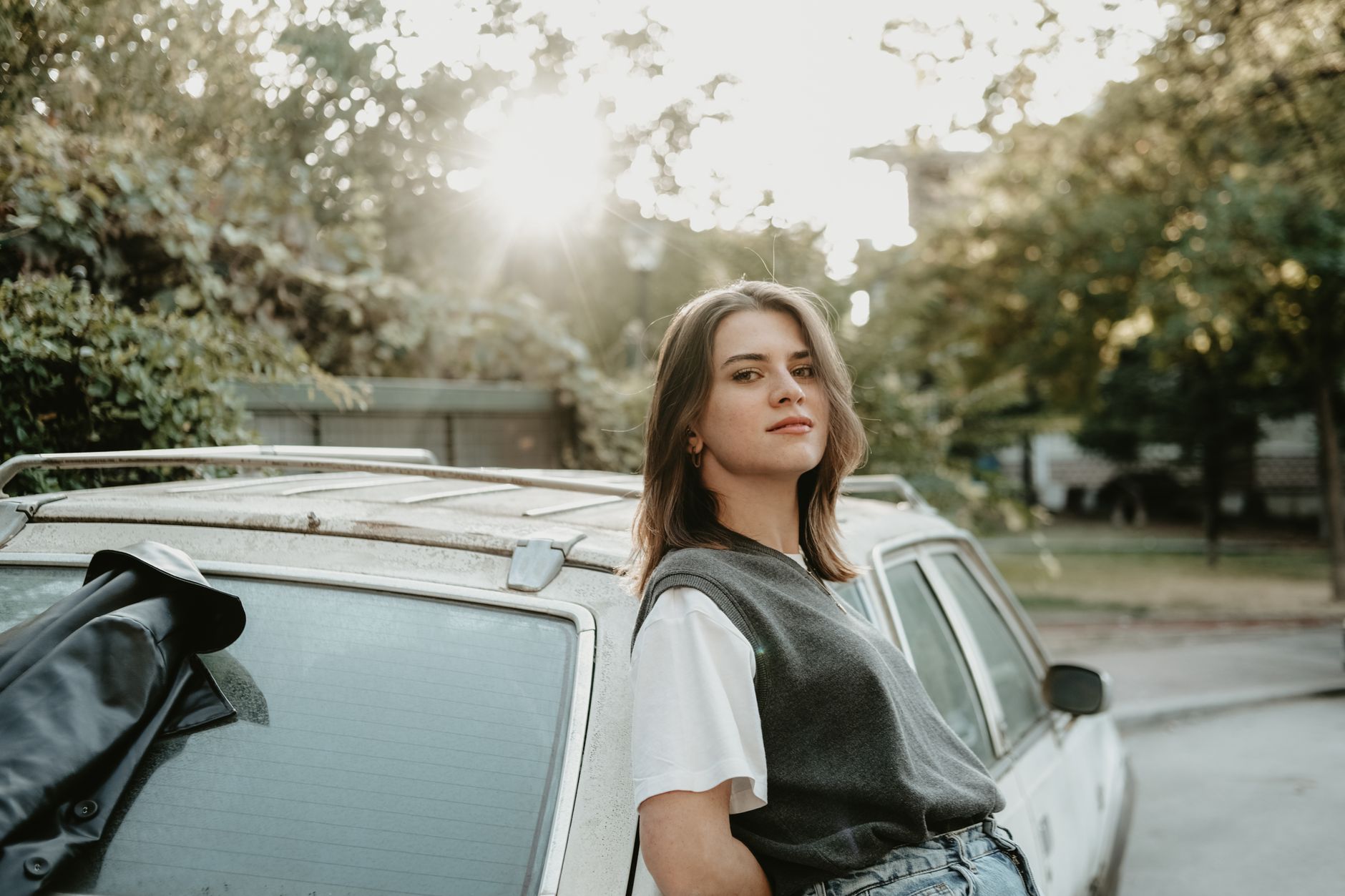A confident young woman leaning on a car, enjoying the sunlight outdoors.