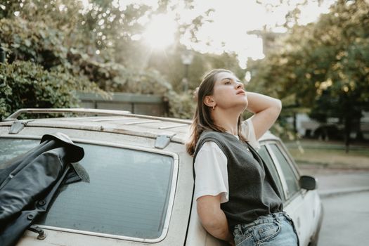 A woman in casual attire leans against a vintage car, basking in sunlight amidst greenery.