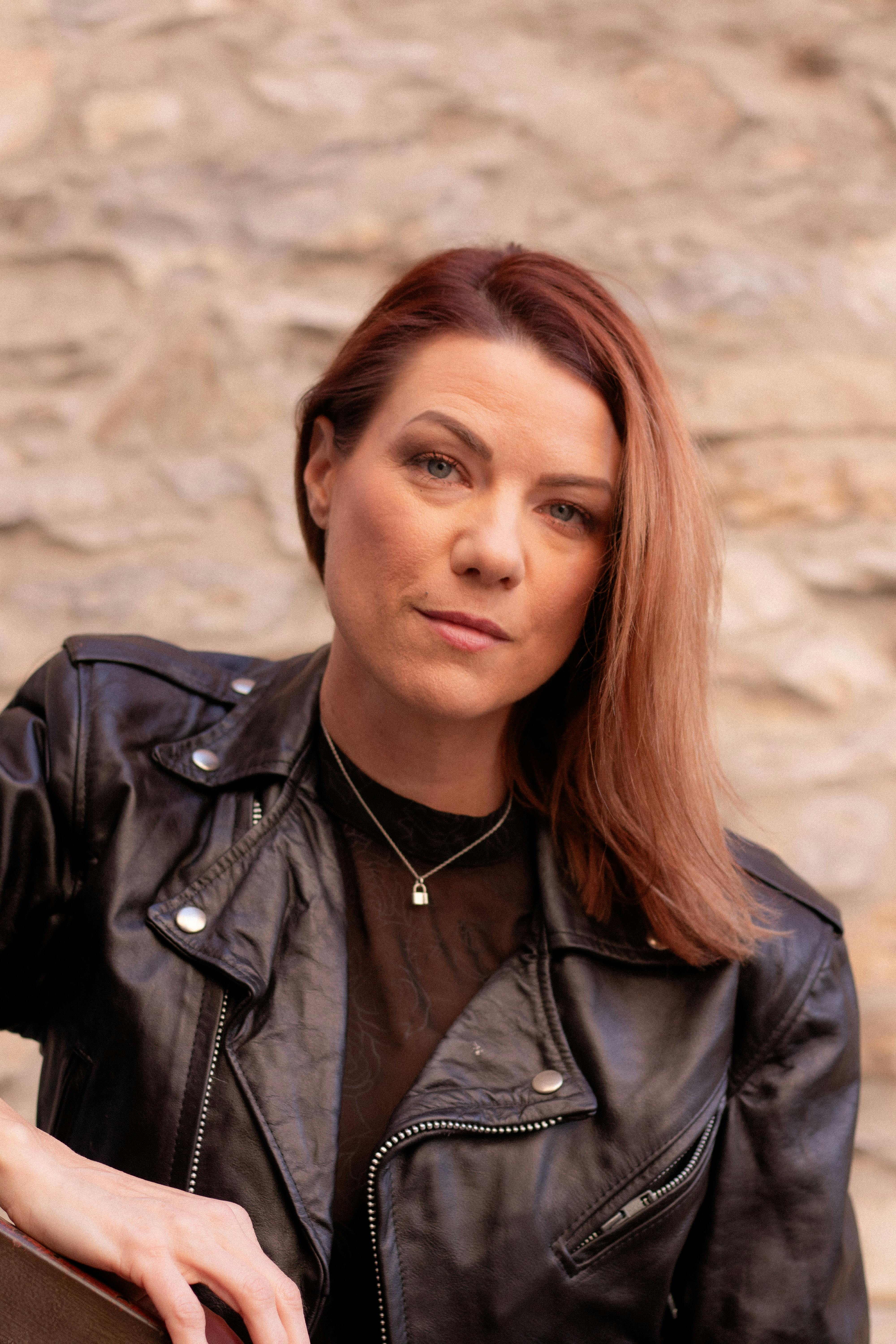 Portrait of a woman in a black leather jacket posing against a rustic stone wall in natural light.
