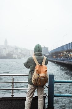 A traveler stands by a riverside railing, gazing at an urban skyline on a cloudy day.