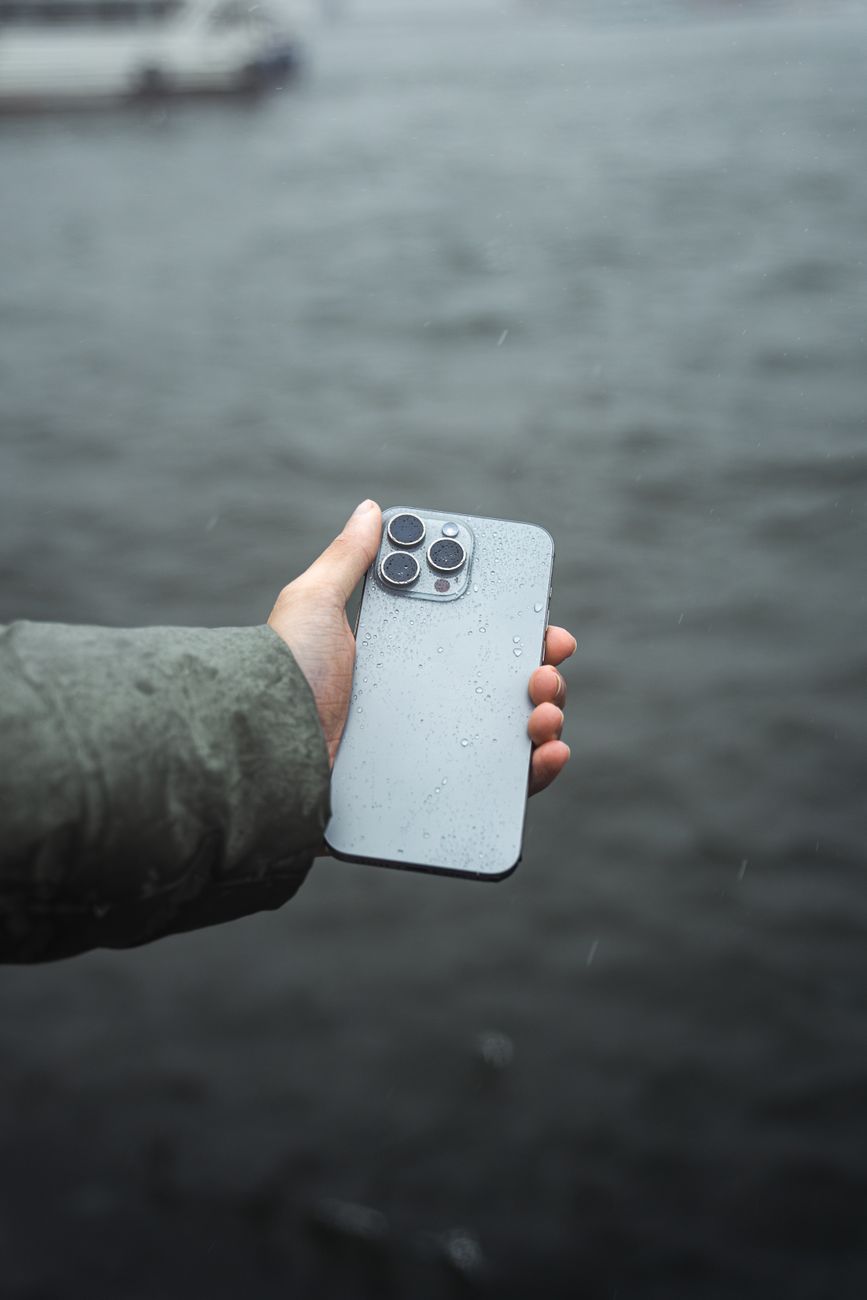 A close-up of a hand holding a modern smartphone with raindrops by a river, showcasing technology in wet conditions.