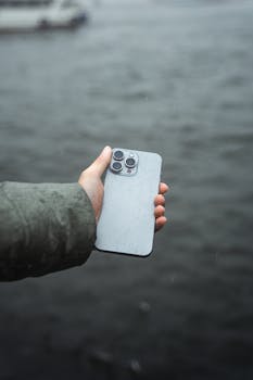 A close-up of a hand holding a modern smartphone with raindrops by a river, showcasing technology in wet conditions.