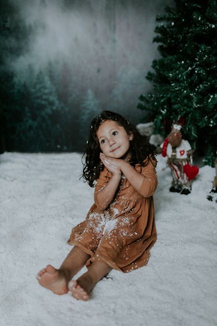 Cute little girl sitting in snow with Christmas tree, capturing festive winter vibes.