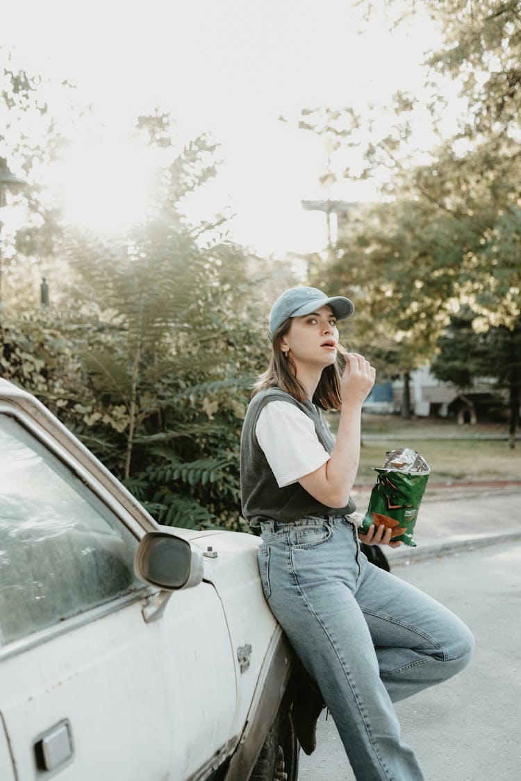 Young Woman Leaning Against A Car And Eating Crisps 