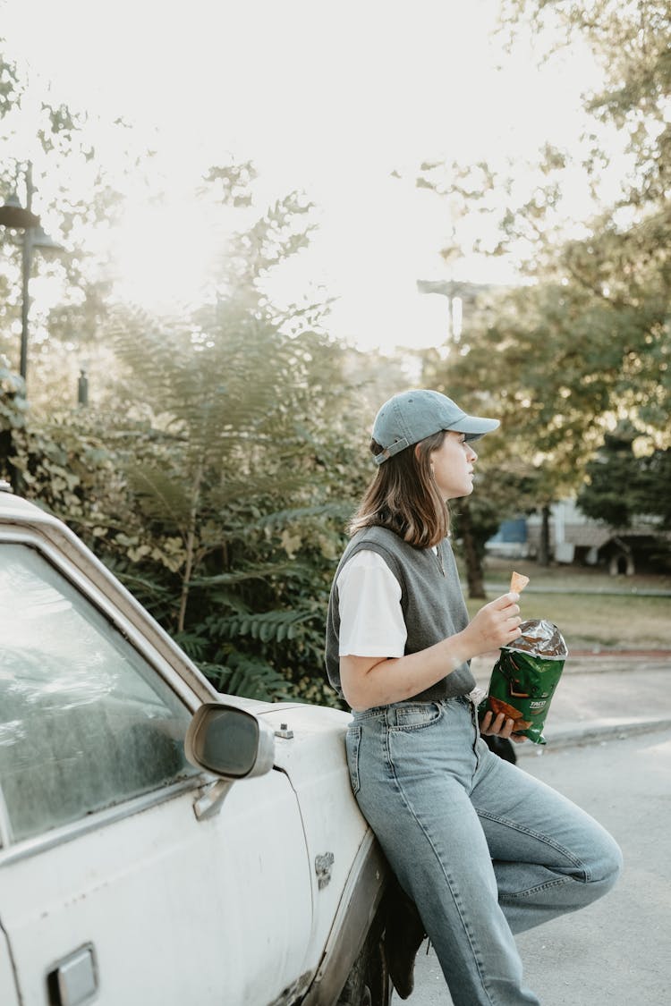 Young Woman Leaning Against A Car And Eating Crisps 