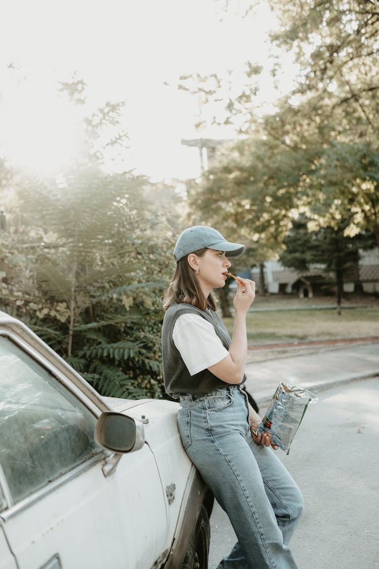 Young Woman Leaning Against A Car And Eating Crisps 