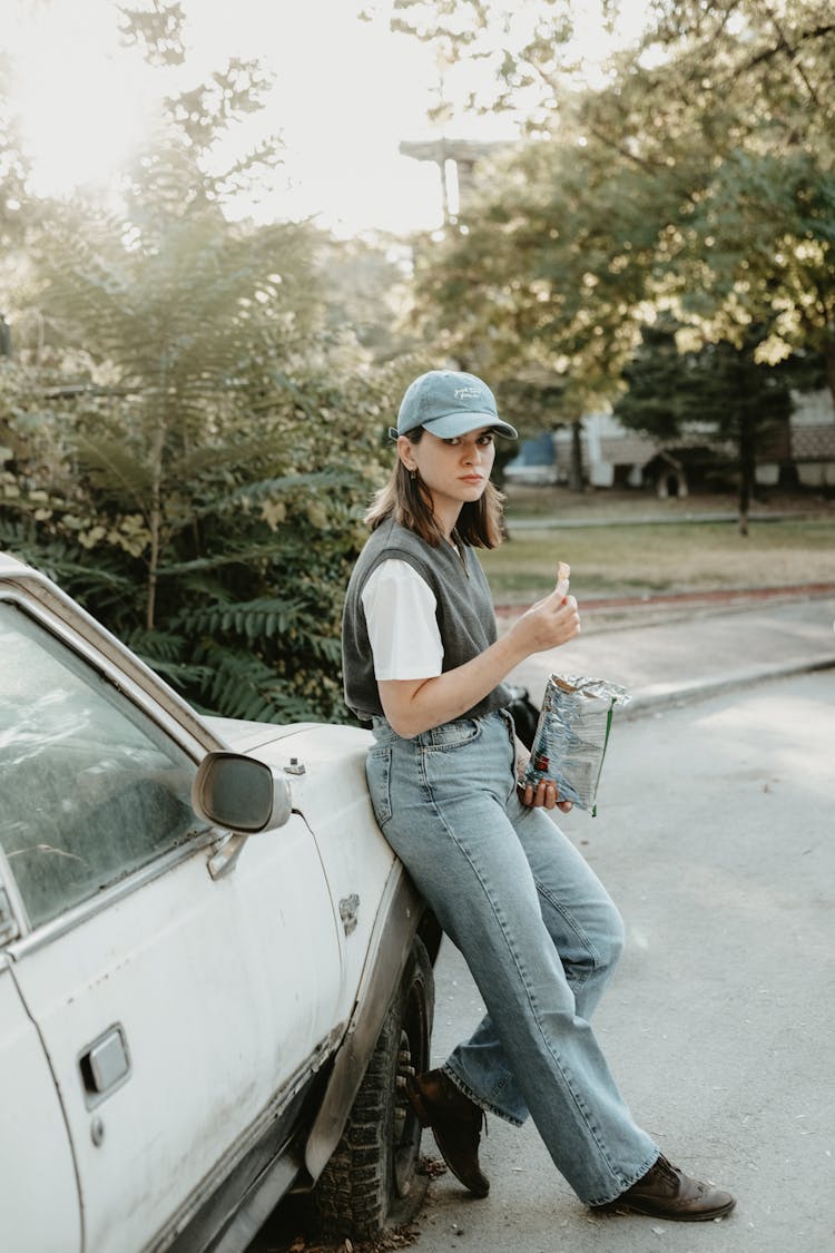 Young Woman Leaning Against A Car And Eating Crisps 