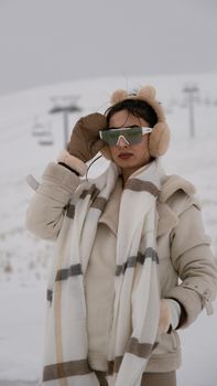 Stylish woman in winter attire with earmuffs and sunglasses on a snowy slope.