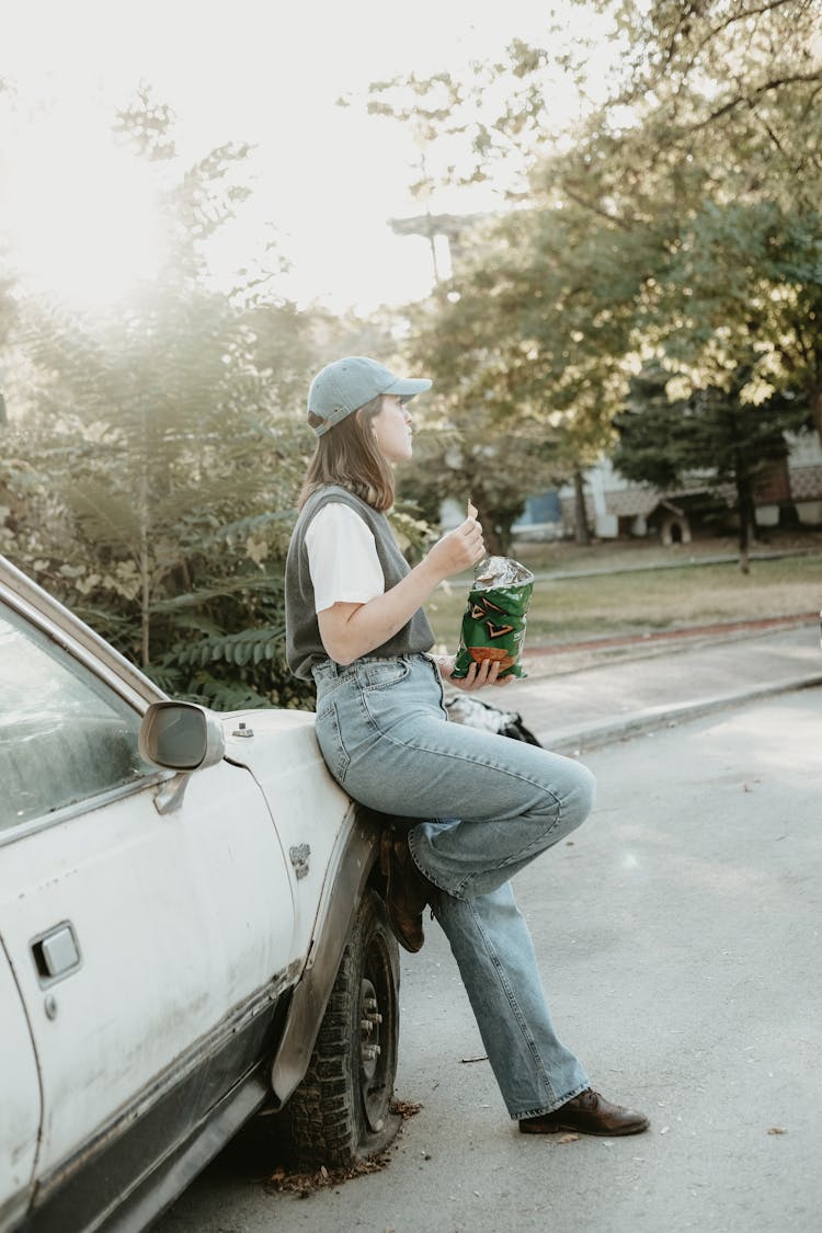 Young Woman Leaning Against A Car And Eating Crisps 