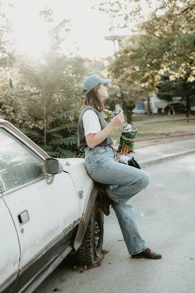 Young Woman Leaning Against A Car And Eating Crisps 