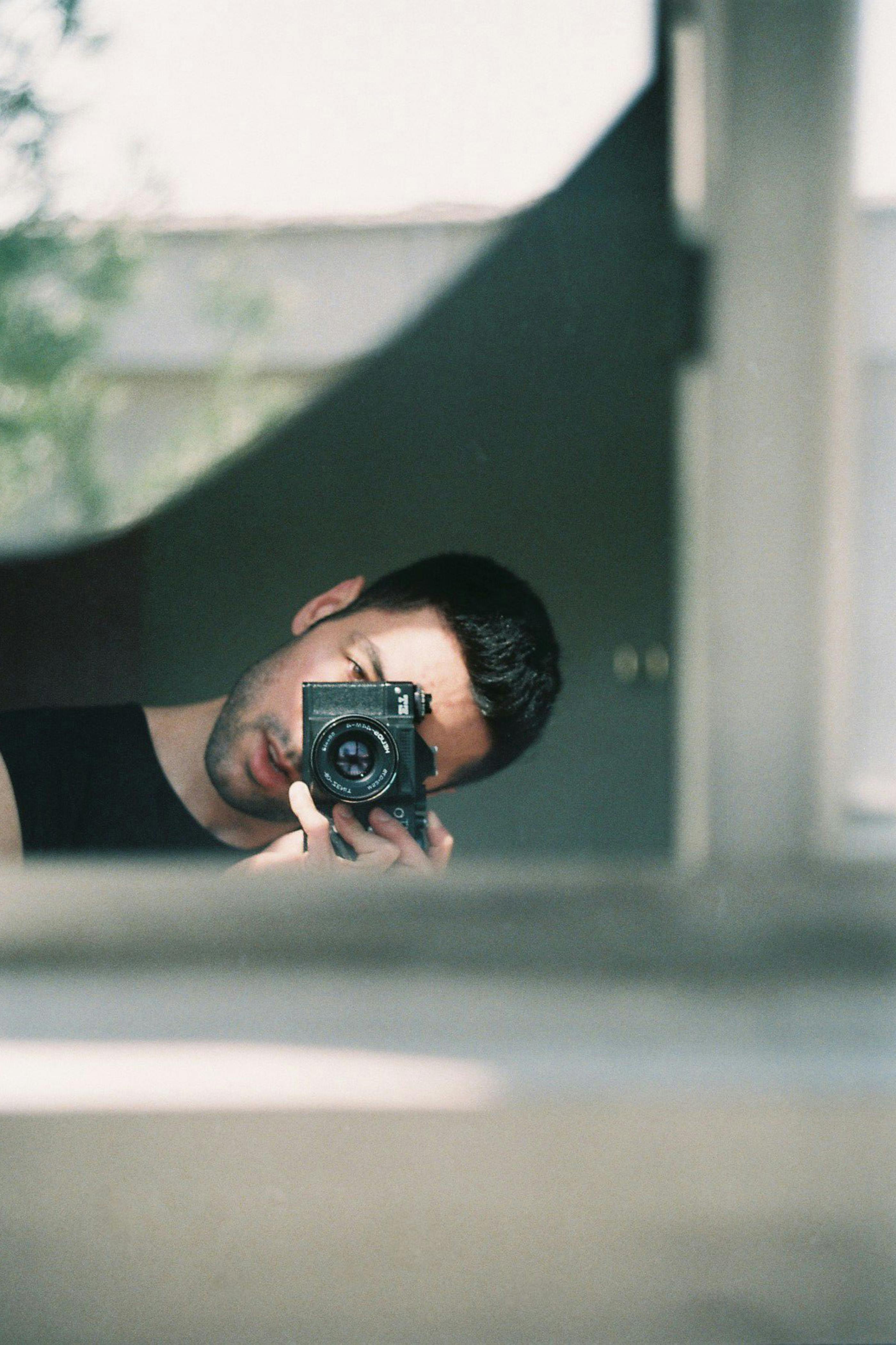 A man takes a selfie with a camera, reflecting in a window with soft natural light.