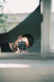 A man takes a selfie with a camera, reflecting in a window with soft natural light.