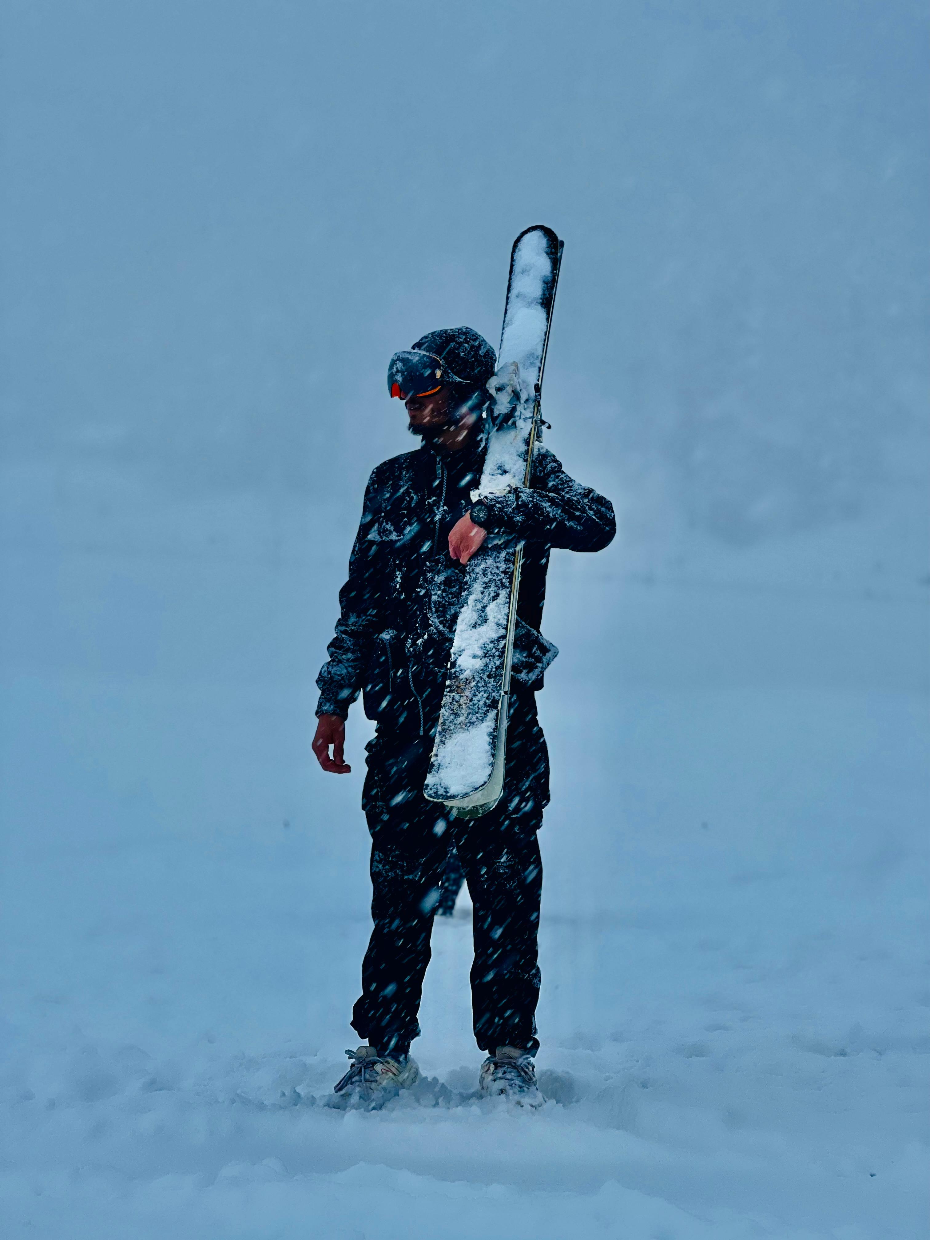 A skier stands in a snowy landscape, holding skis in winter gear with snow falling.