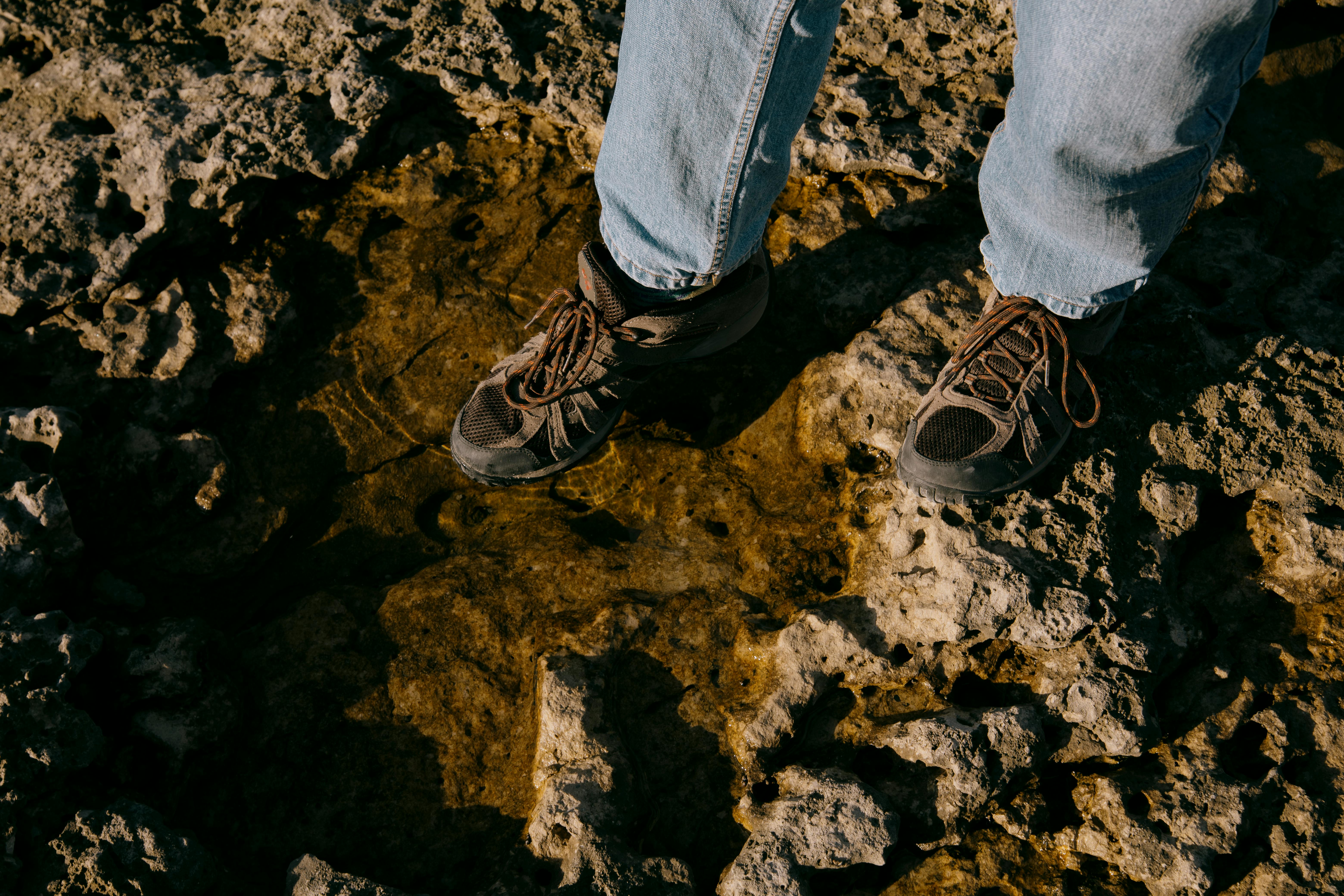 A hiker standing on rocky ground with water puddles, wearing casual shoes and jeans.