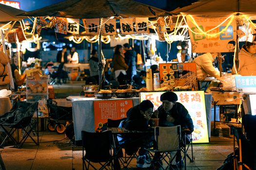 Bustling night market scene with food stalls illuminated and patrons enjoying street food under warm lights.