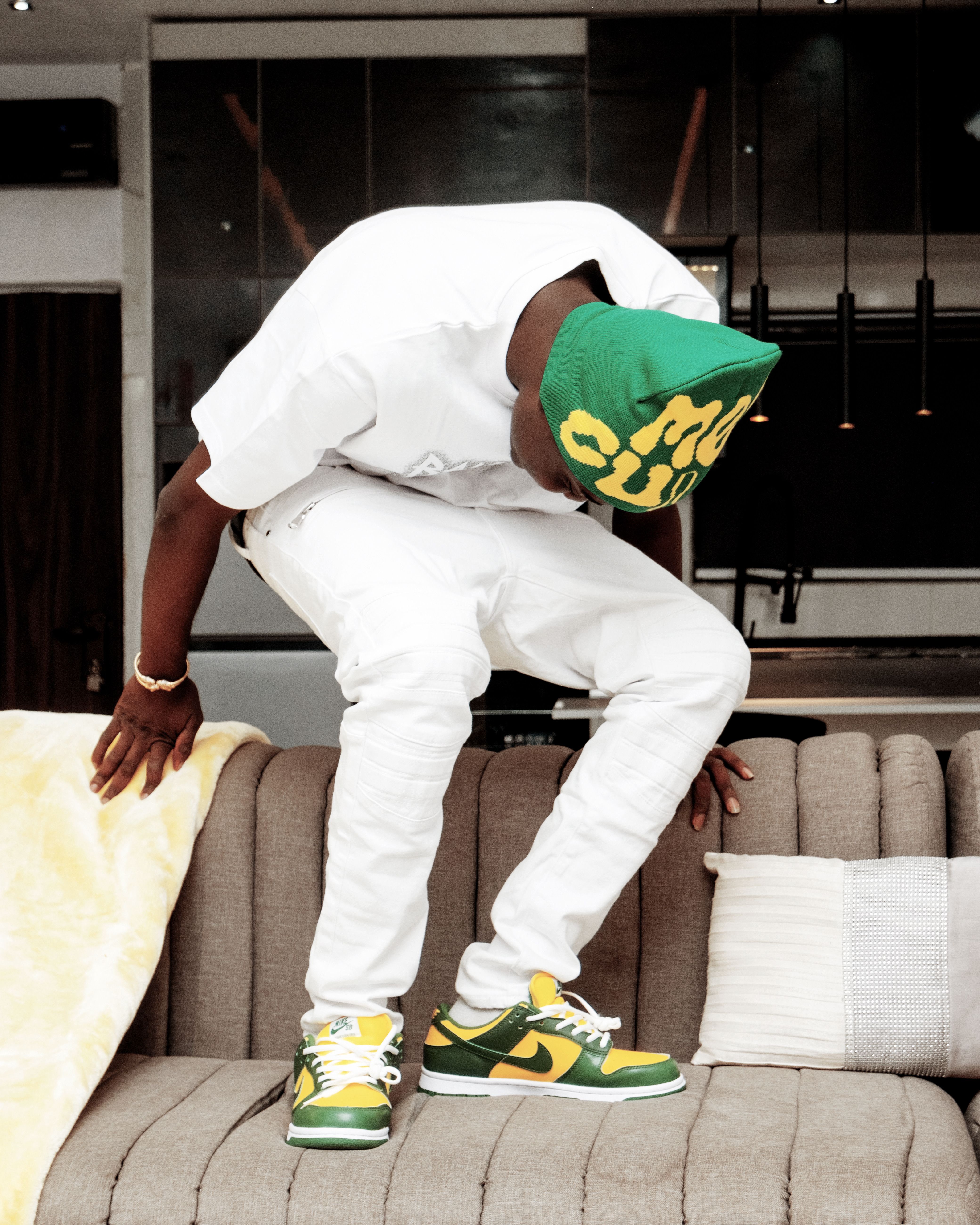 A fashionable man in white outfit and green cap posing on a sofa indoors. Stylish urban vibe.