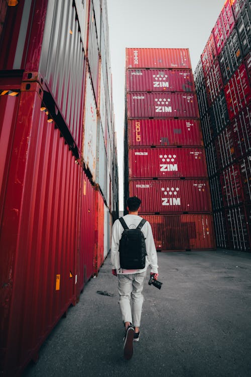 Man with Backpack Walking among Red Containers at Warehouse