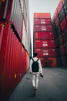 A man walks with a backpack and camera, surrounded by towering red cargo containers in Brisbane's logistics area.
