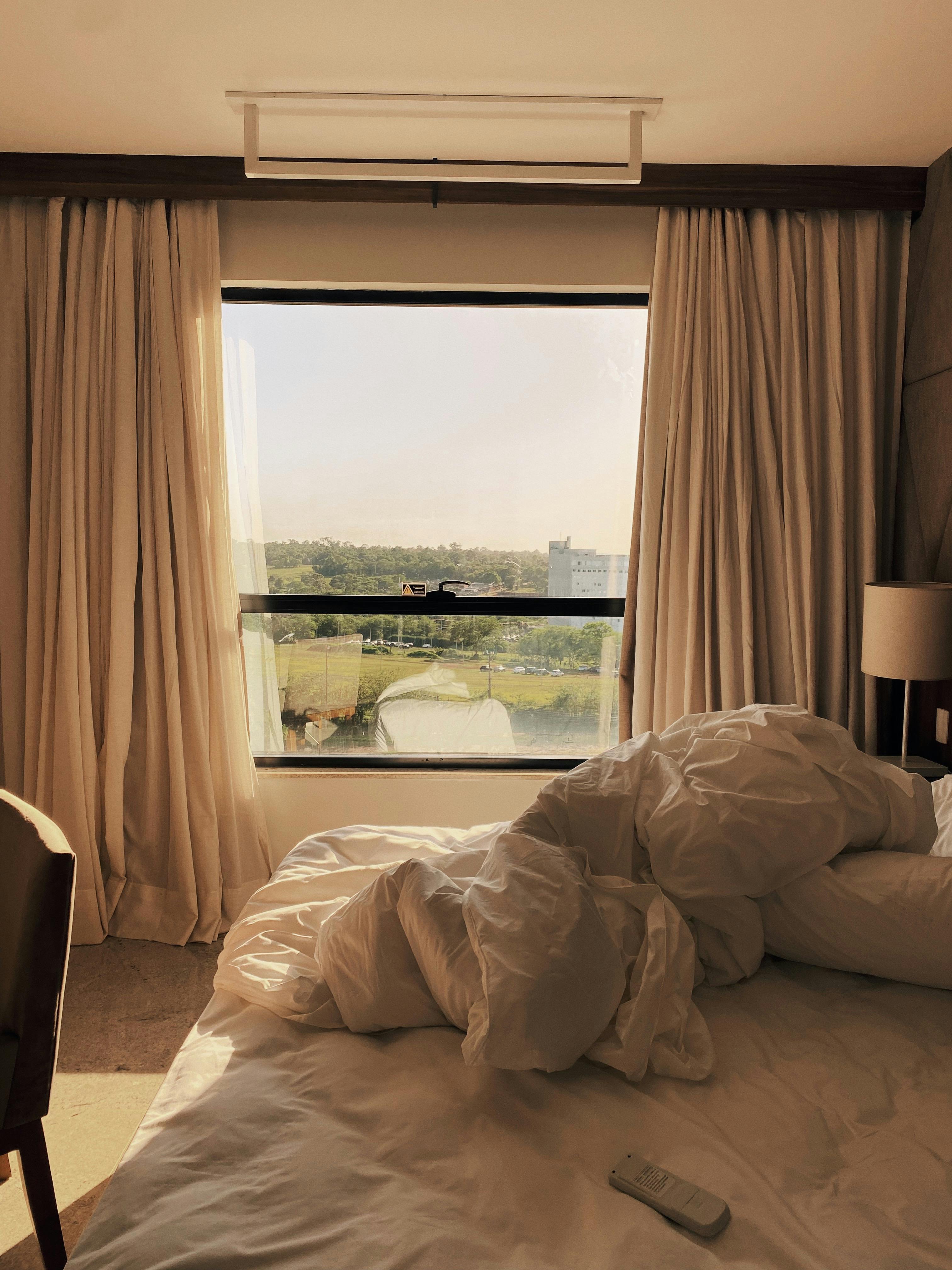 Sunlit hotel room with unmade bed and scenic window view for travel comfort.