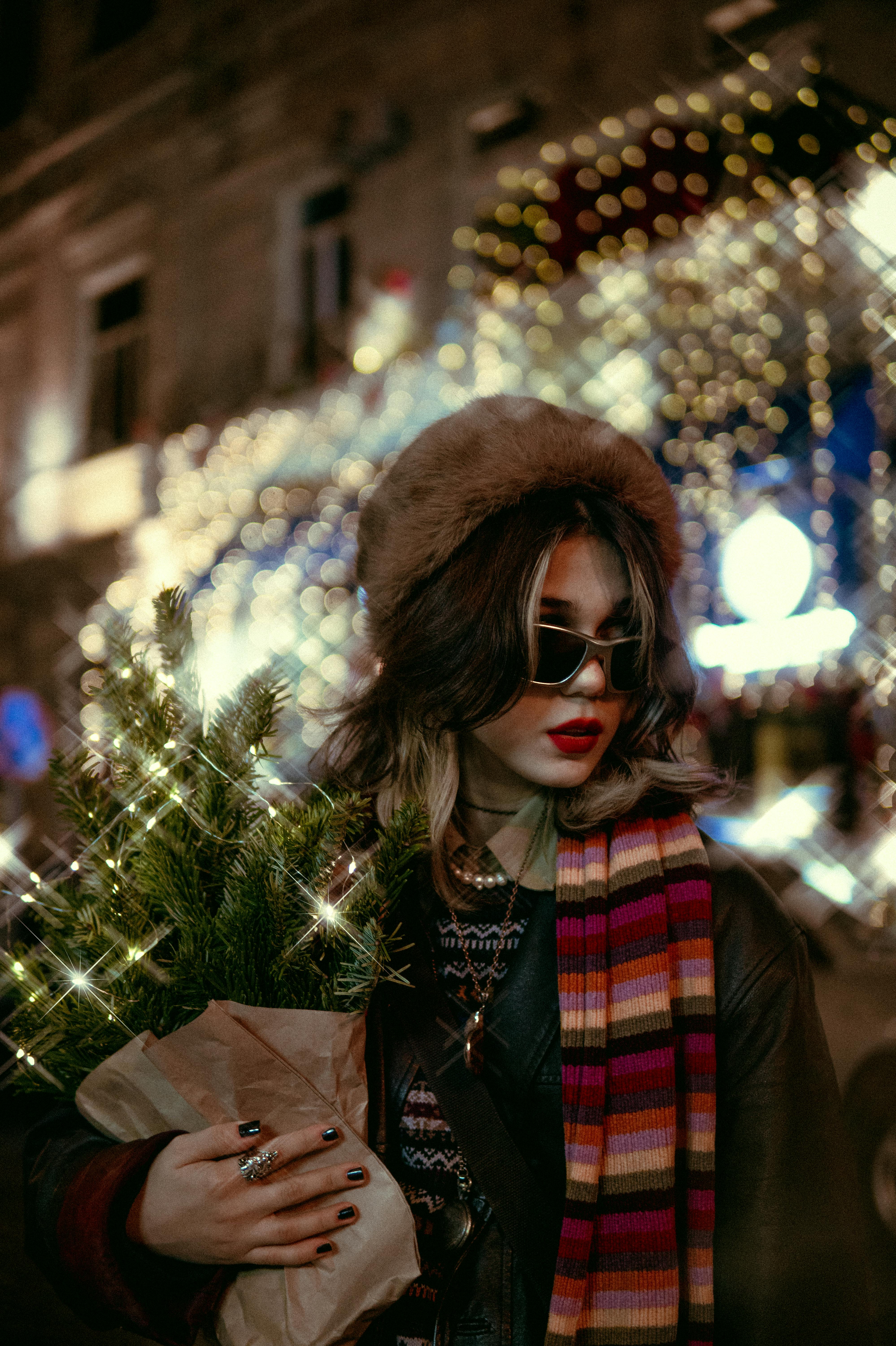 Fashionable woman with sunglasses and fur hat in Christmas-lit city street holding a small tree.