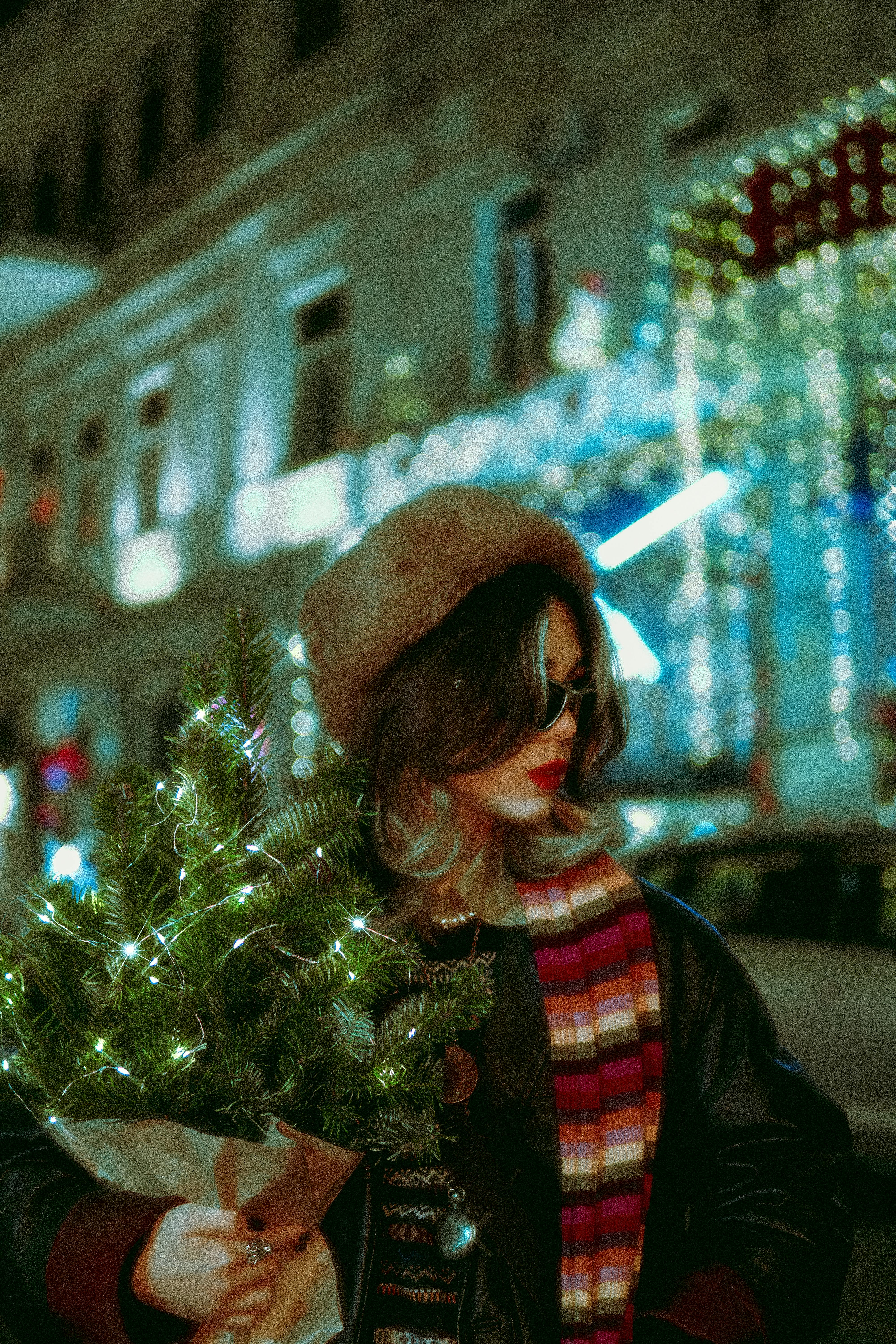 Fashionable woman holding a Christmas tree in a vibrant urban setting at night.