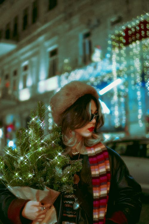 Woman in Hat and Scarf and with Christmas Branches at Night