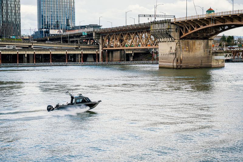 A motorboat speeds on the river under a bridge in Portland, OR. Urban skyline in backdrop.