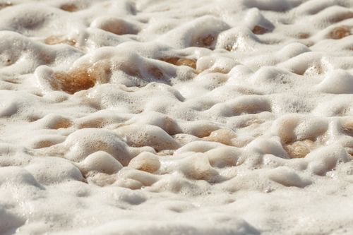 Free Detailed view of cascading ocean foam forming patterns on a sandy beach under sunlight. Stock Photo