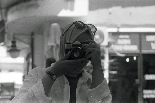 Free Black and white photo of a woman capturing urban street scene with an analog camera. Stock Photo