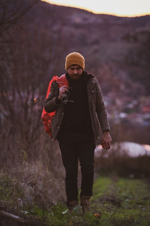 Free A man carrying a red backpack walks in a mountainous area at sunset, wearing a yellow hat. Stock Photo