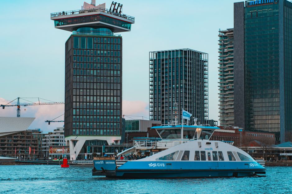 Ferry in front of iconic A'DAM Tower and modern skyscrapers in Amsterdam harbor.