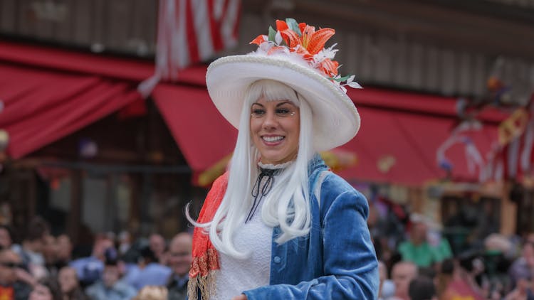 Woman Walking In The Parades In New York, Wearing A White Hat Adorned With US Flags, White Hair, And A Red And Blue Dress