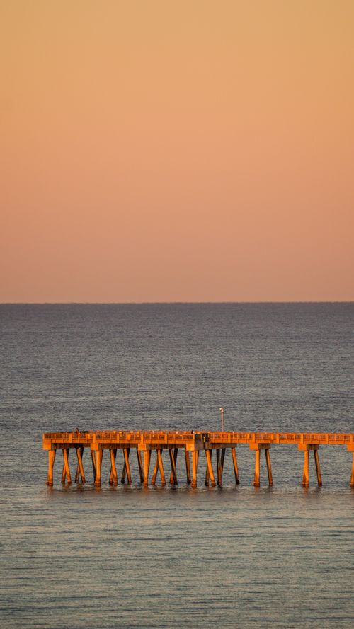 Free A serene view of a pier extending over a calm ocean at sunset, captured in warm tones. Stock Photo