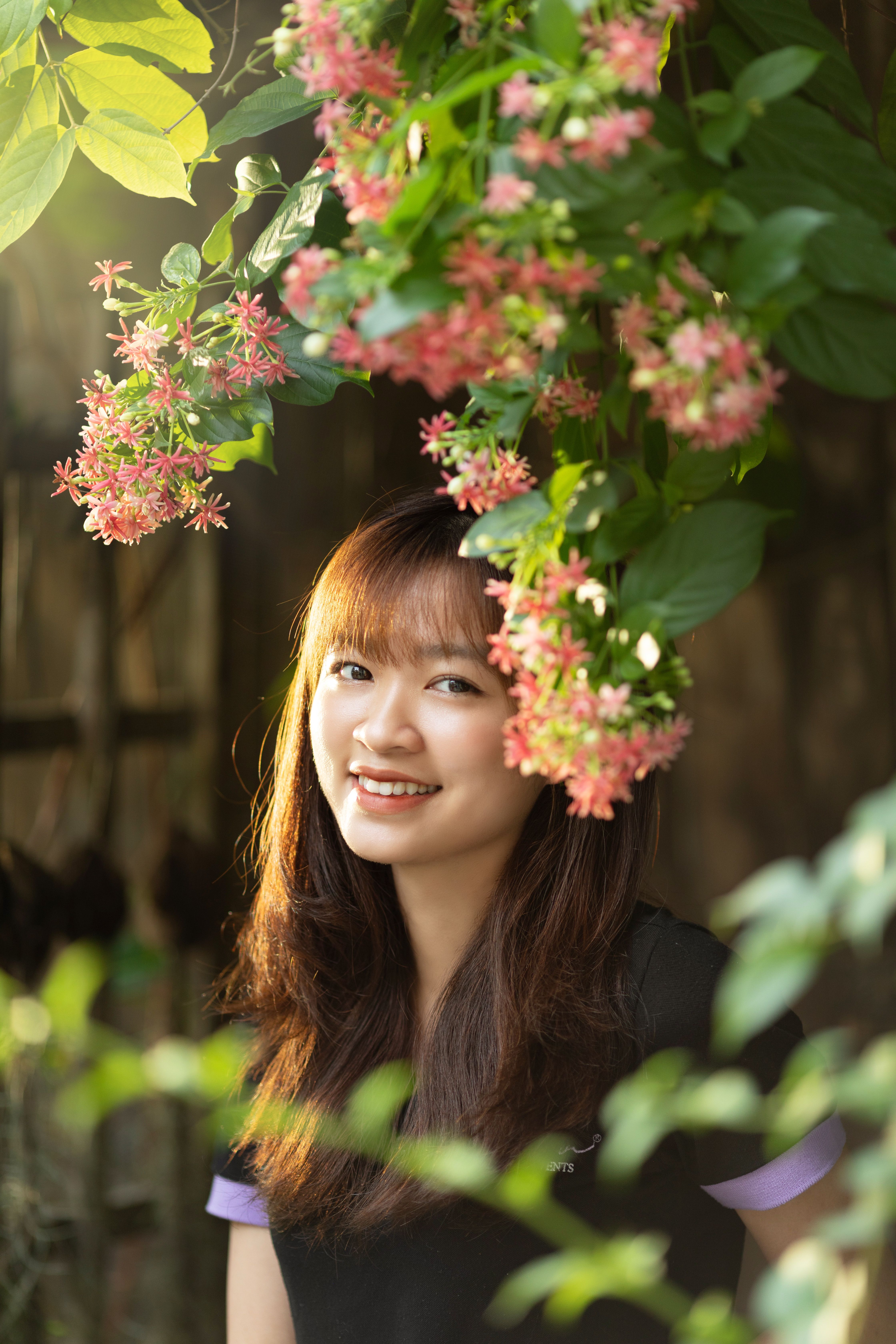 A woman smiles surrounded by vibrant pink flowers in a sunlit garden.