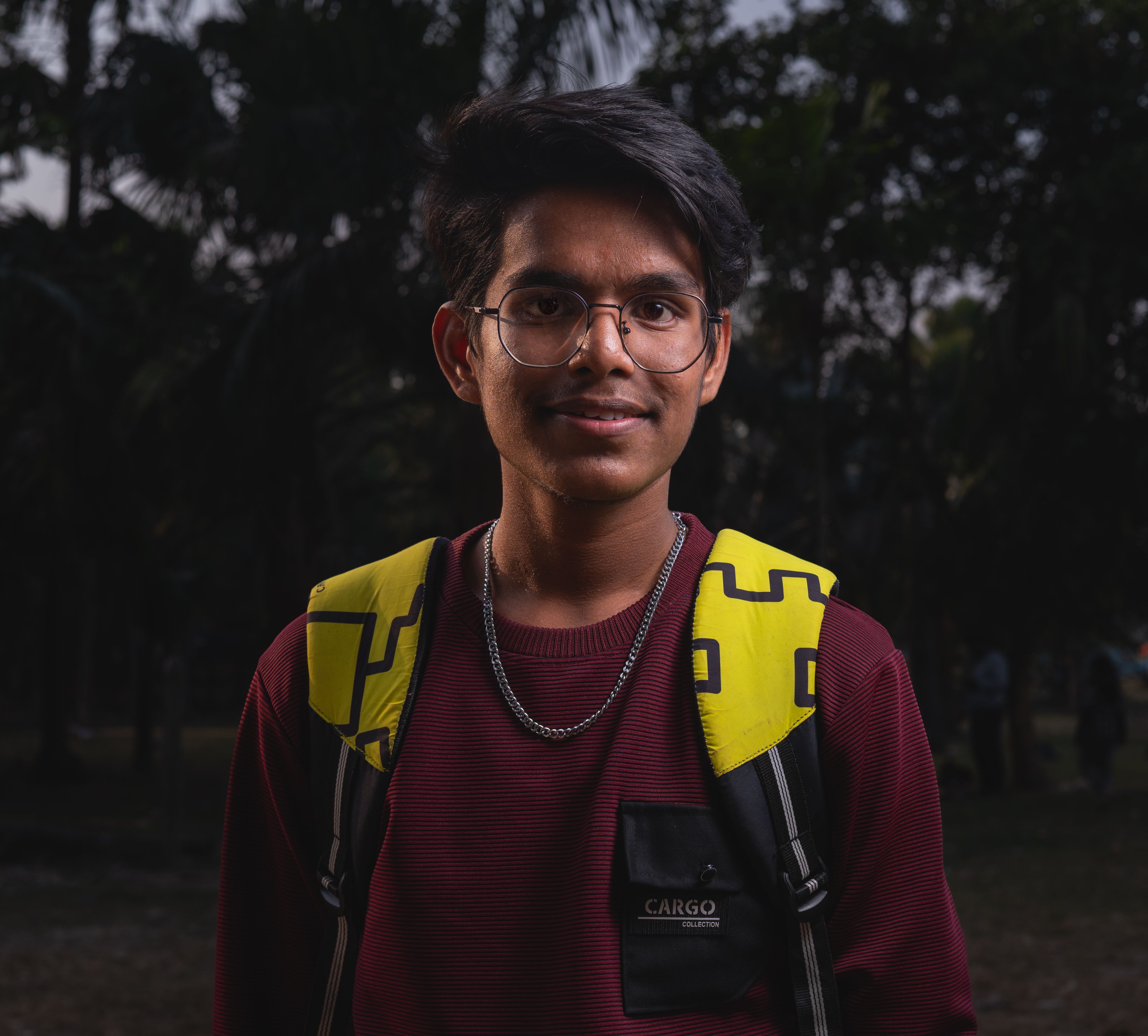 Portrait of a young man wearing a backpack and glasses, smiling outdoors in a dark, wooded area.