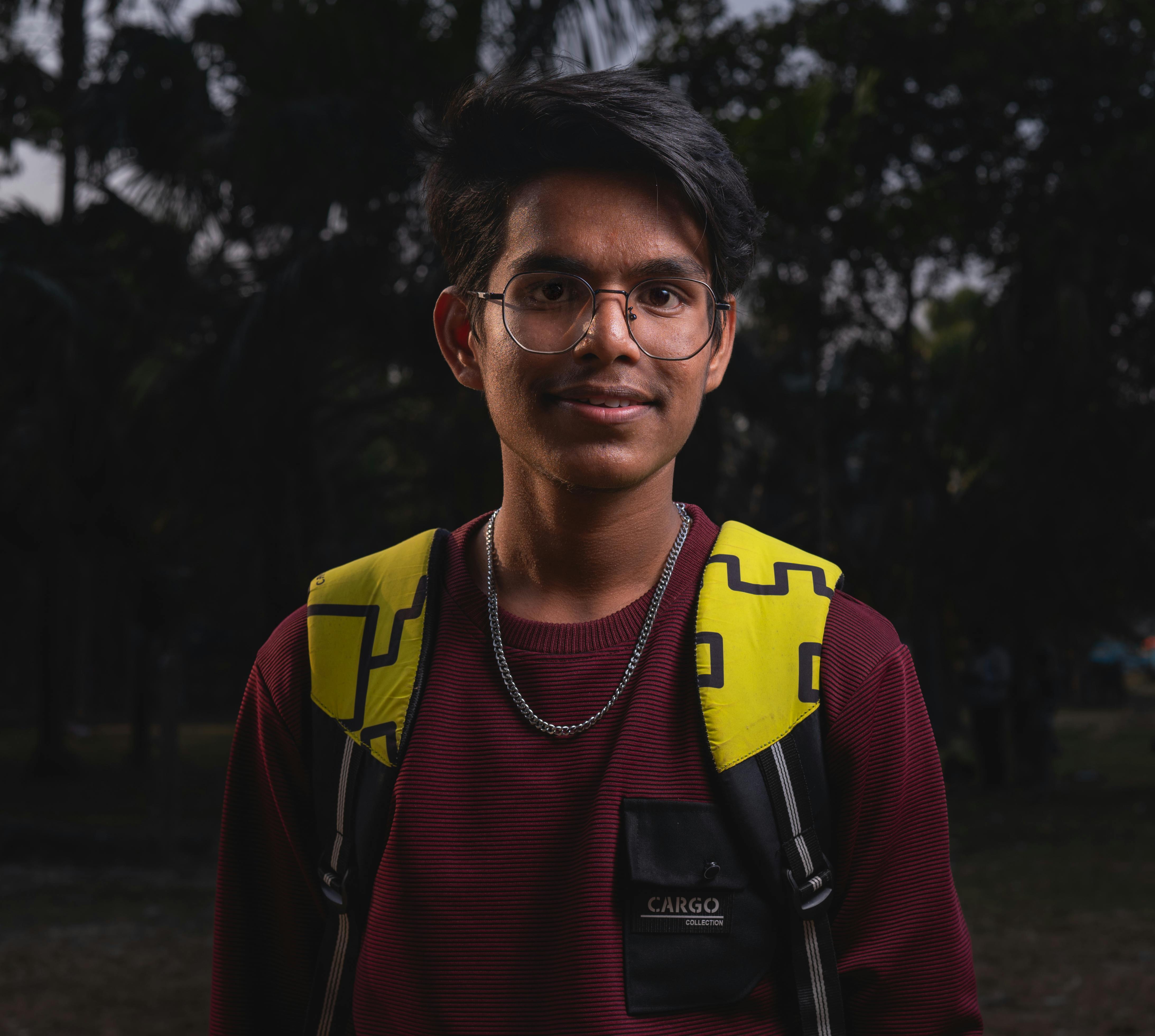 A young adult wearing eyeglasses and a backpack standing outdoors in a park during twilight.