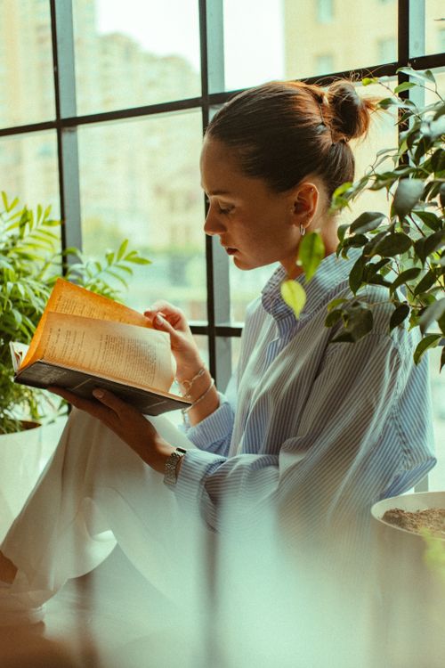 Free Woman reading a book by a window surrounded by indoor plants, enjoying a serene moment. Stock Photo