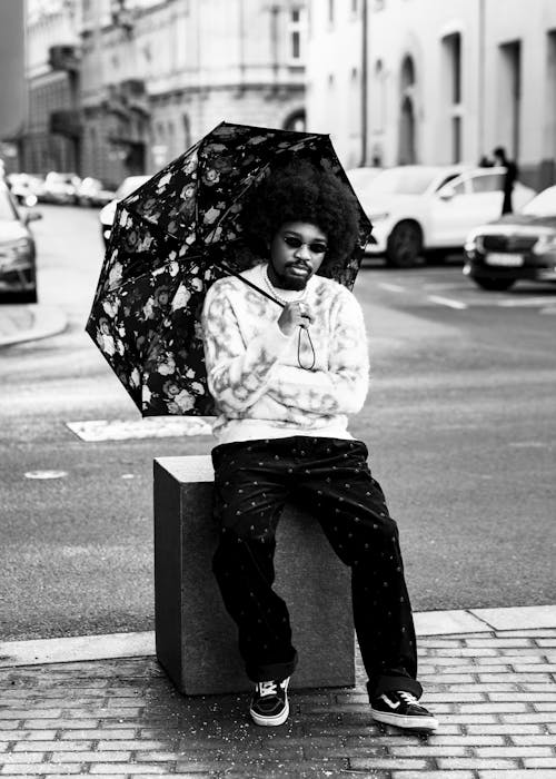 Man in a Trendy Outfit Sitting on a Bollard on a Sidewalk and Holding an Umbrella 