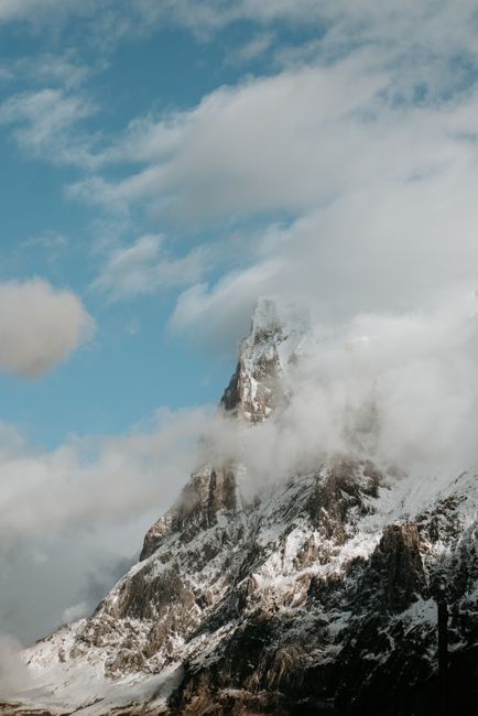 A breathtaking view of a snow-capped mountain peak partially covered by clouds, reaching into a clear blue sky.