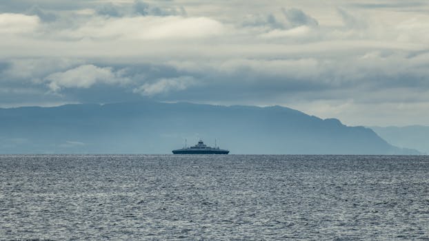 Distant ferry sailing across a calm sea under cloudy skies with mountain backdrop.