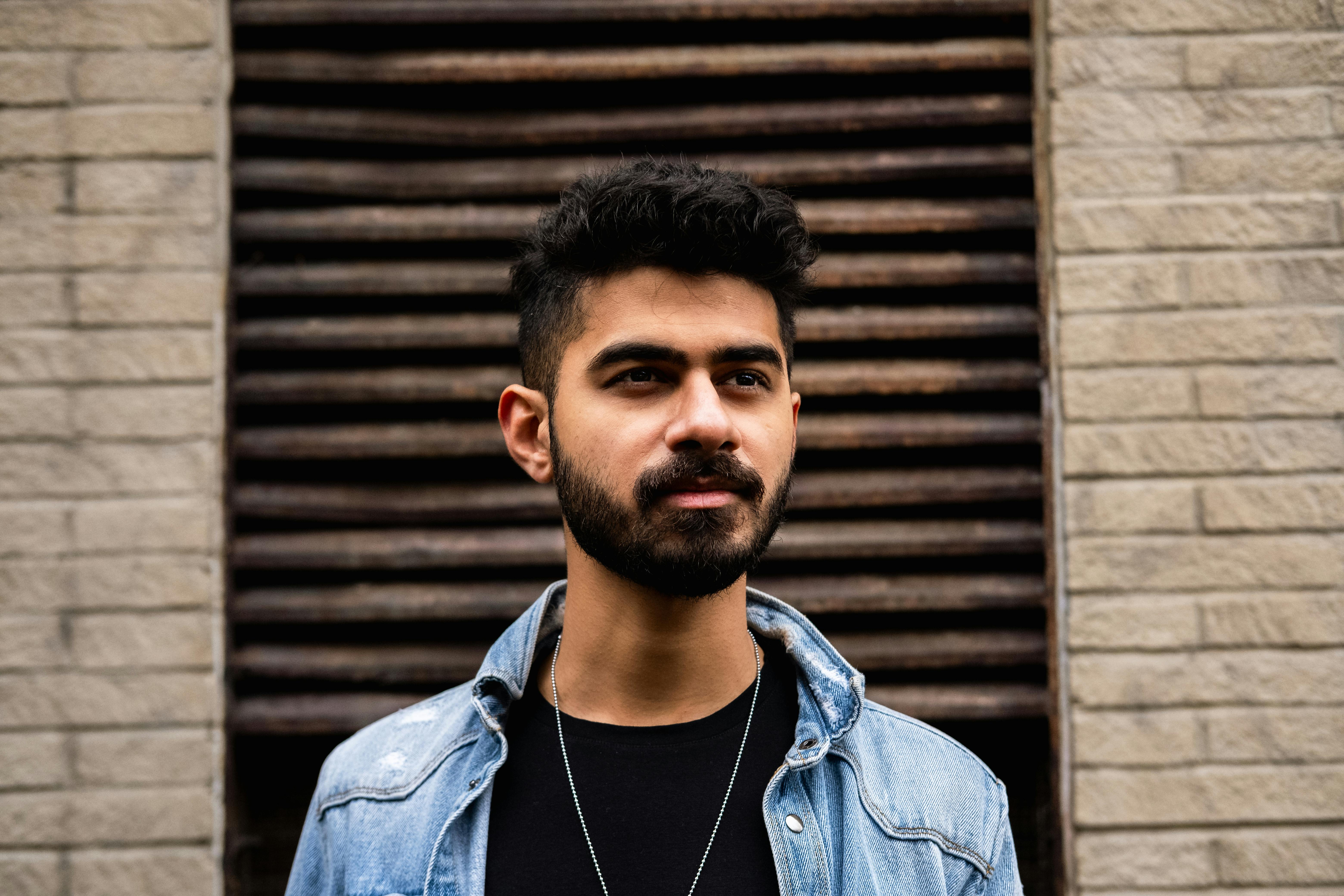 A young bearded man with dark hair wearing a denim jacket, standing outdoors against a textured brick wall.