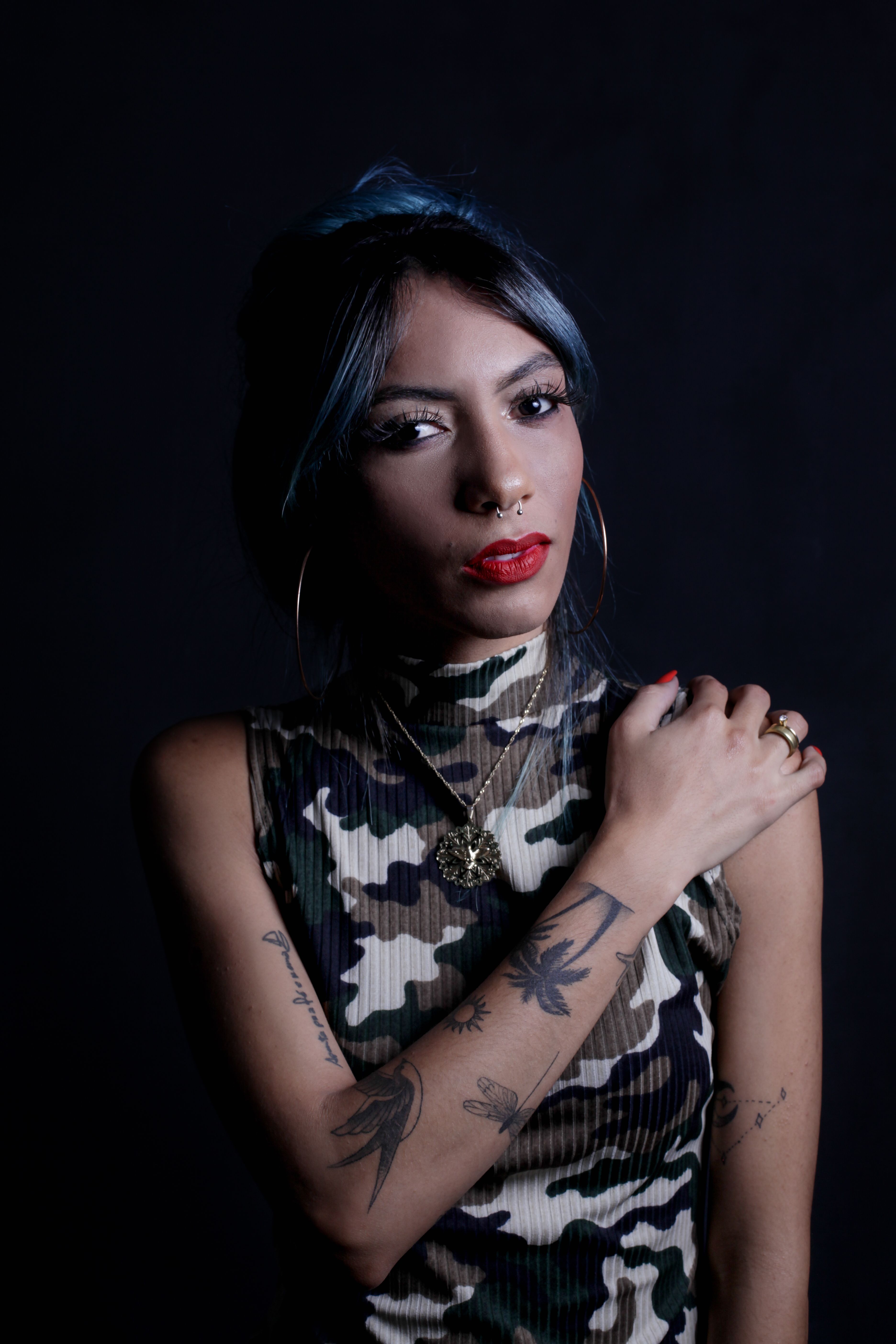 Studio portrait of a woman with tattoos and red lips in camouflage attire against a black backdrop.