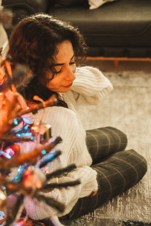 Brunette Woman in White Sweater and Gray Pants Sitting under a Christmas Tree